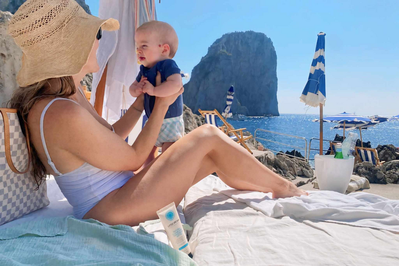 Woman holding baby at a sunny beach with blue umbrellas and a rocky sea backdrop