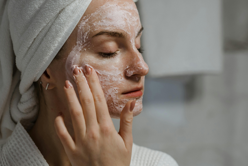 Woman applying a cream face mask with eyes closed, wearing a towel and bathrobe